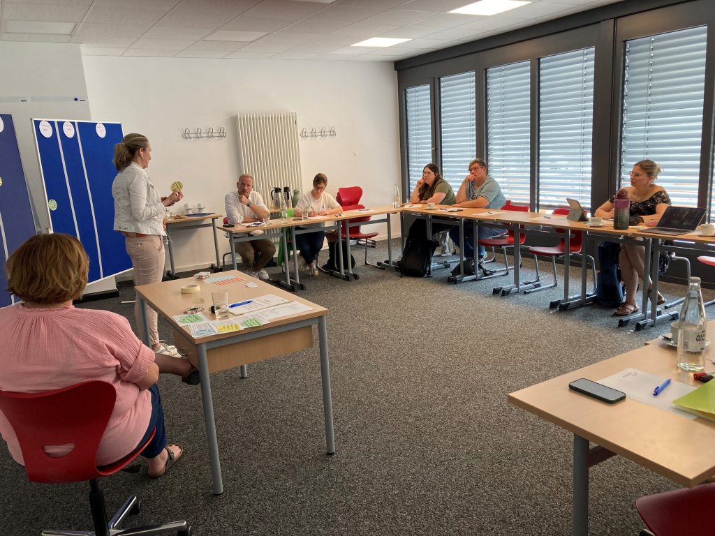 Verschiedene Personen sitzen in einem Halbkreis an Tischen und hören einer Person zu