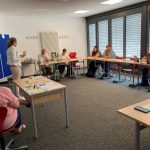 Verschiedene Personen sitzen in einem Halbkreis an Tischen und hören einer Person zu
